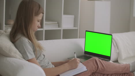 school-girl-is-viewing-online-lesson-on-laptop-with-green-screen-for-chroma-key-technologywriting-at-exercise-book-learning-from-home